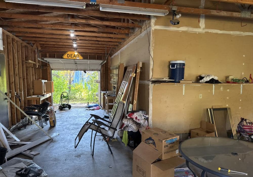 Cluttered garage interior with storage boxes, chairs, and backyard view through open door.